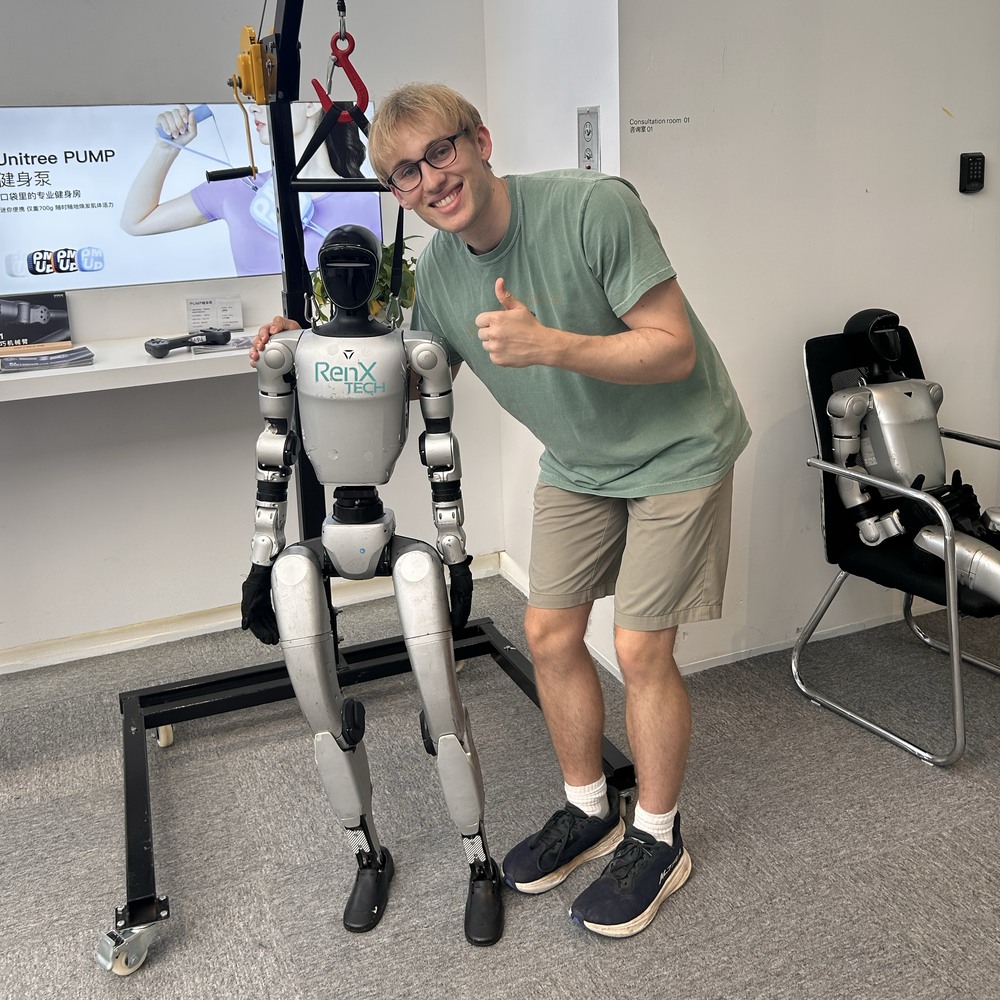 Will smiling beside a standing humanoid robot, with his hand on its shoulder.